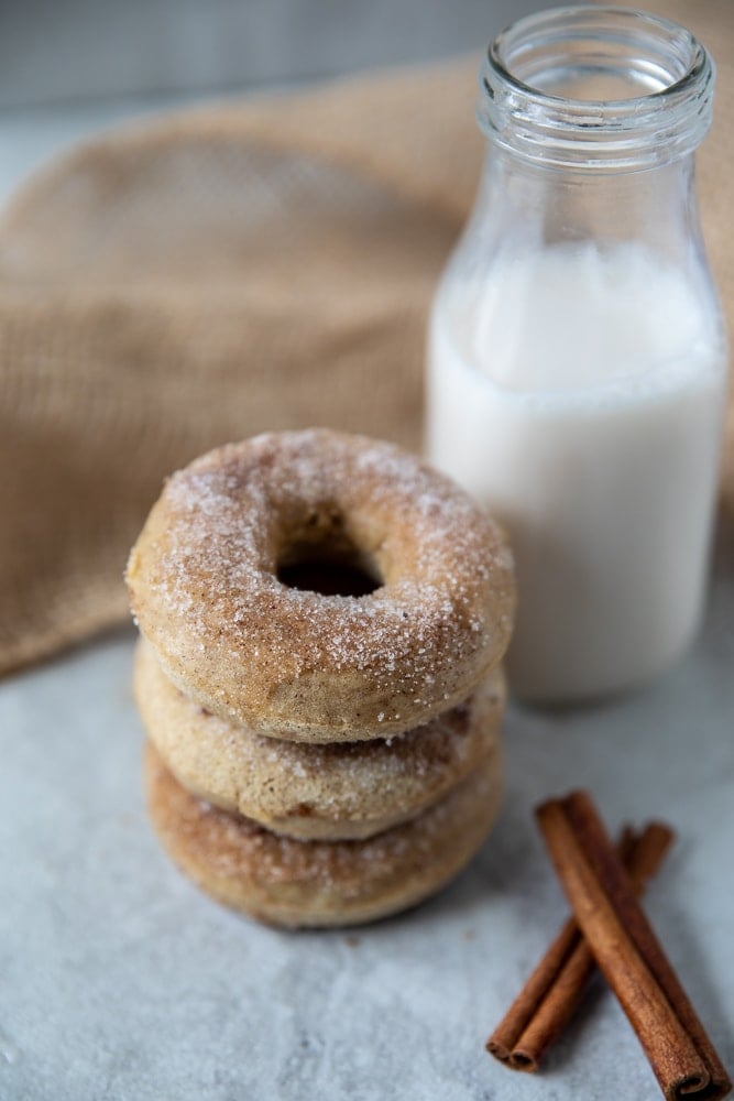3 Cinnamon donuts stacked up next to cinnamon sticks and a glass of milk. 