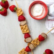 Waffles, breakfast sausage, and strawberries on a stick next to yogurt