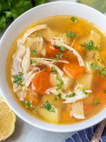 Chicken soup with potatoes and carrots in white bowl next to lemon