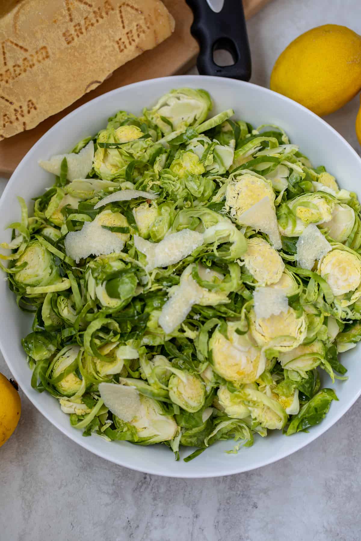 Shaved Brussels Sprouts Salad in White Bowl next to parmesan and lemons.