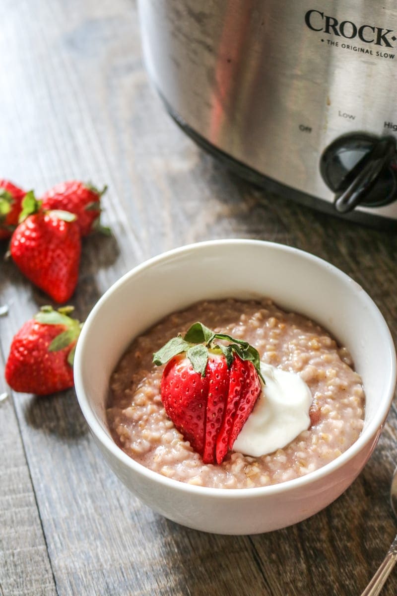 Bowl of overnight steel cut oatmeal in a white bowl with strawberries and yogurt next to crock pot