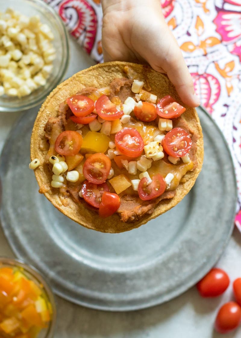 Child's hand holding a baked tostada topped with tomatoes and corn.