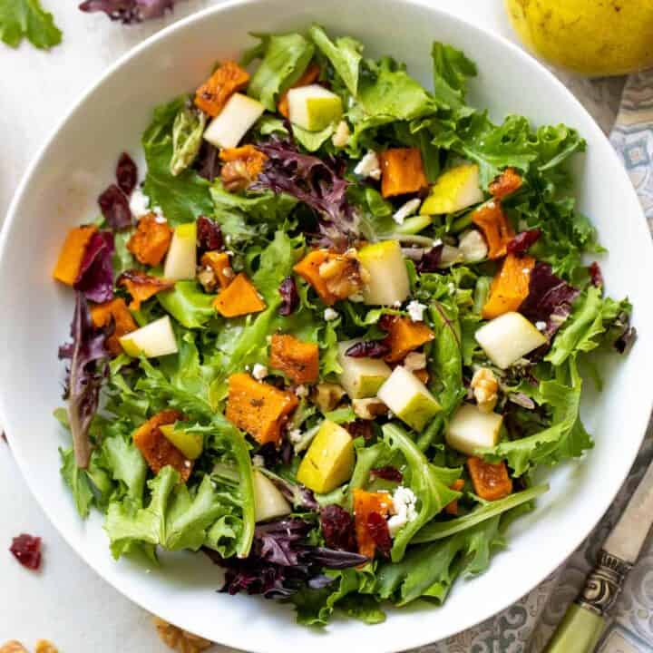 A blue pottery bowl filled with baby mixed greens, roasted butternut squash, pear cubes, feat and dried cranberries.