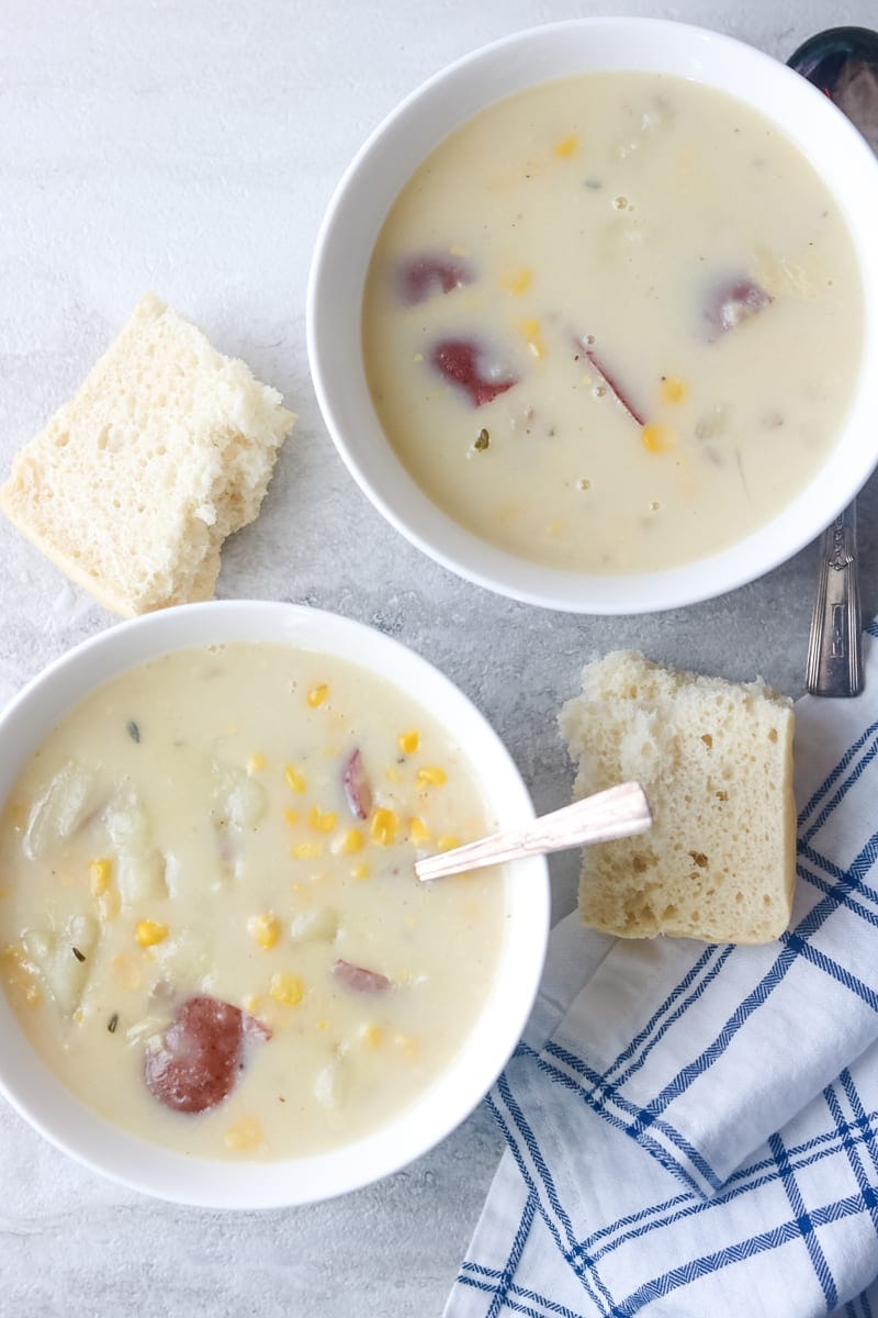 2 white bolws filled with creamy corn and red potato chowder with crusty bread served on the side. 