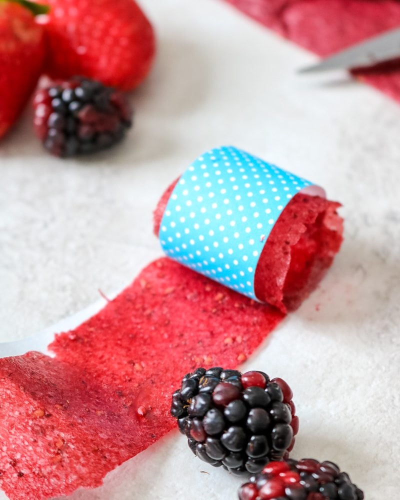 A homemade fruit roll-up rolled up in blue craft paper with blackberries and strawberries to the side.