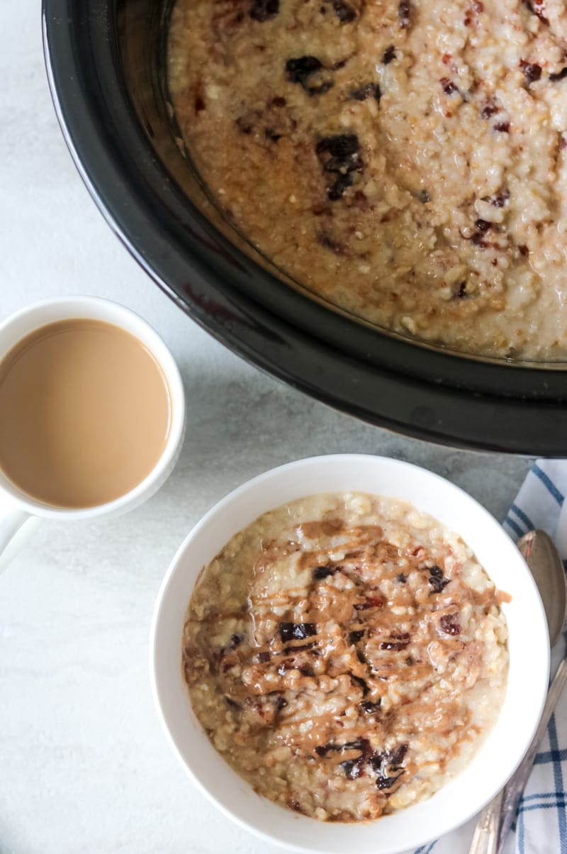 Slow cooker filled with peanut butter and jelly steel cut oatmeal and a bowl of PB&J oatmeal in a white bowl next to coffee.