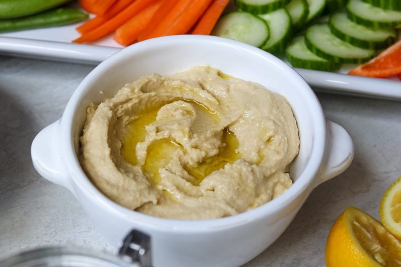 Hummus in white bowl topped with olive oil next to veggies