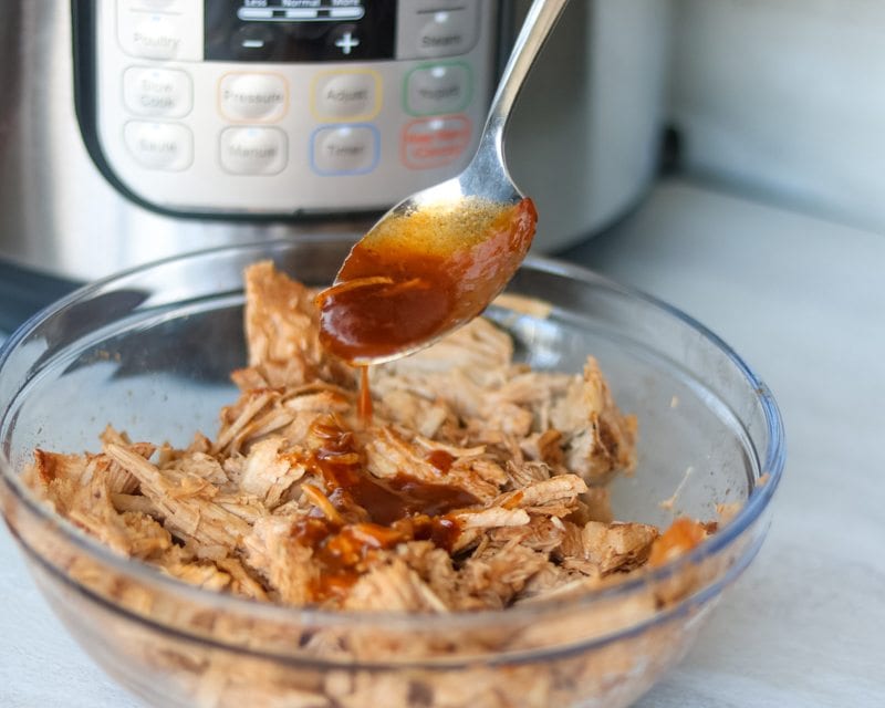 Barbecue sauce being spooned over instant pot shredded pork in bowl with pressure cooker in background. 