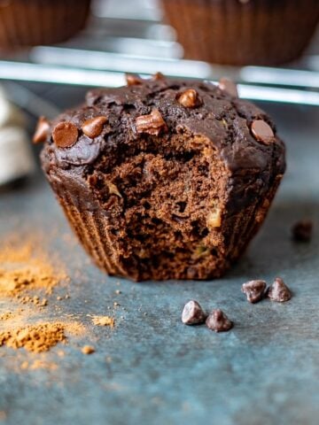 Chocolate Zucchini Muffin with bite out of it next to chocolate chips and cocoa powder