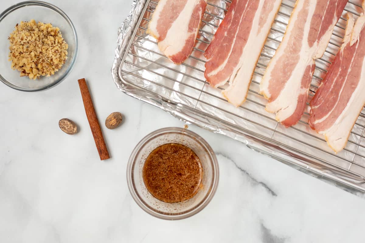 Maple syrup in small bowl with spices next to bacon on sheet pan.