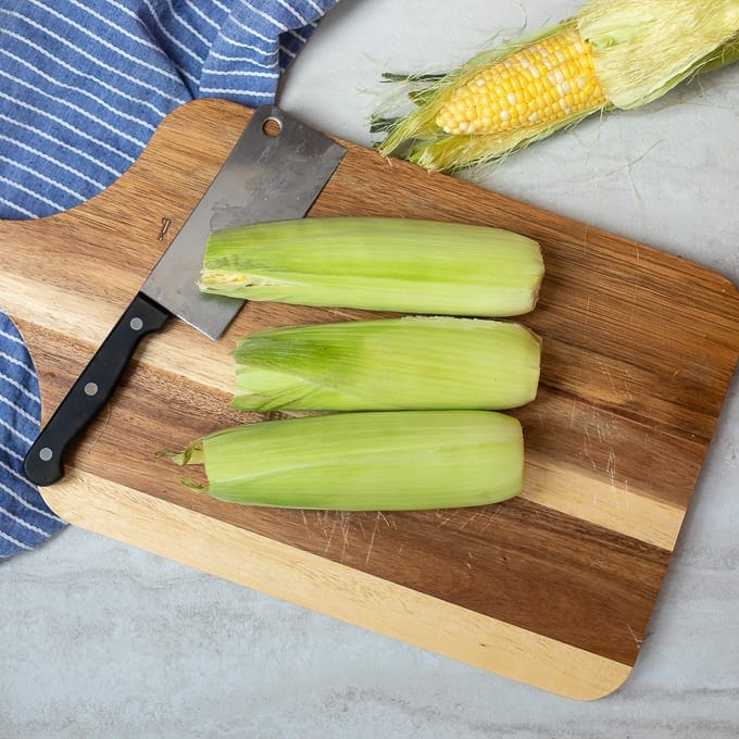 Corn on the cob in husks on wooden cutting board.