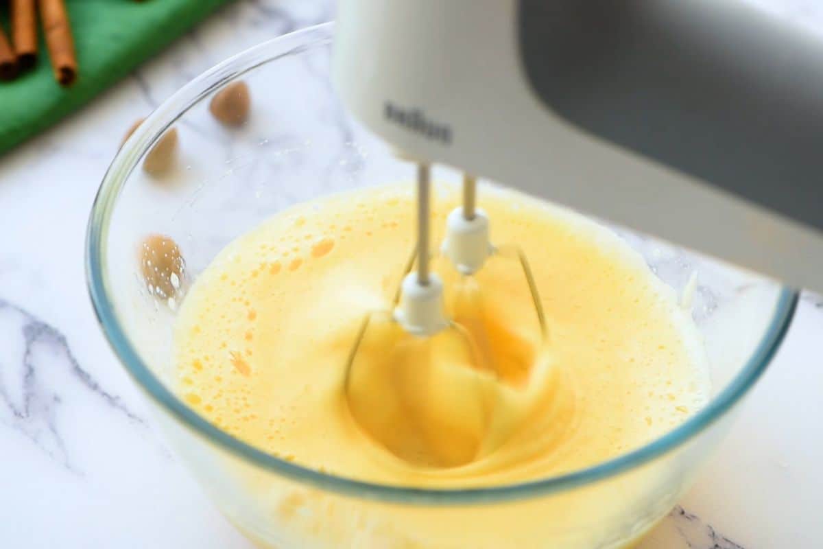 Handheld mixer beating eggs until fluffy in clear mixing bowl.