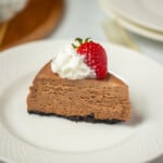 Slice of chocolate cheesecake on white plate topped with whipped cream and strawberry.
