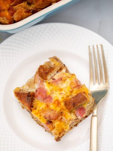 Slice of ham and cheese breakfast casserole on white plate next to fork.