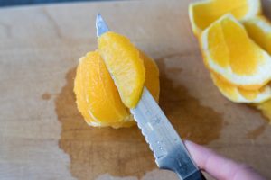 A sharp knife removing an orange section from a peeled orange.