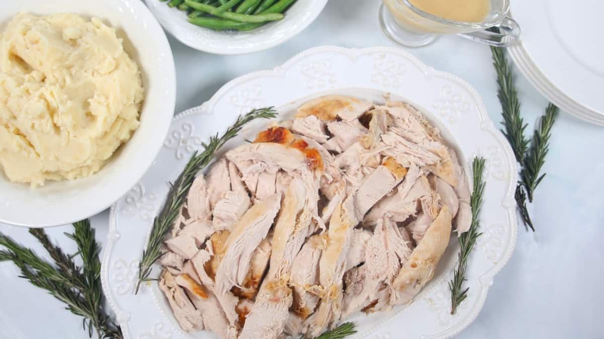 Sliced Turkey Breast on white platter with mashed potatoes and green beans in background.