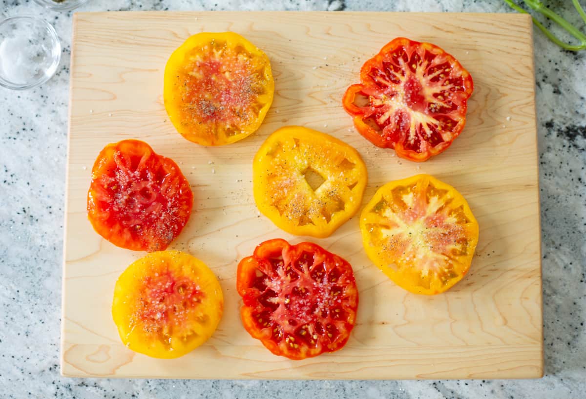 Slices of Heirloom tomatoes on cutting board seasoned with salt and pepper.