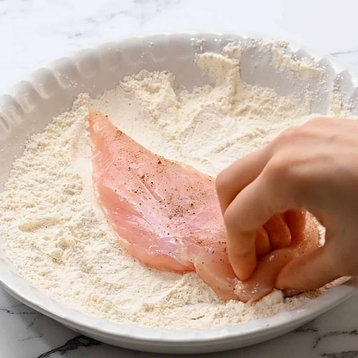 Hand showing dredging a chicken cutlet through seasoned flour.