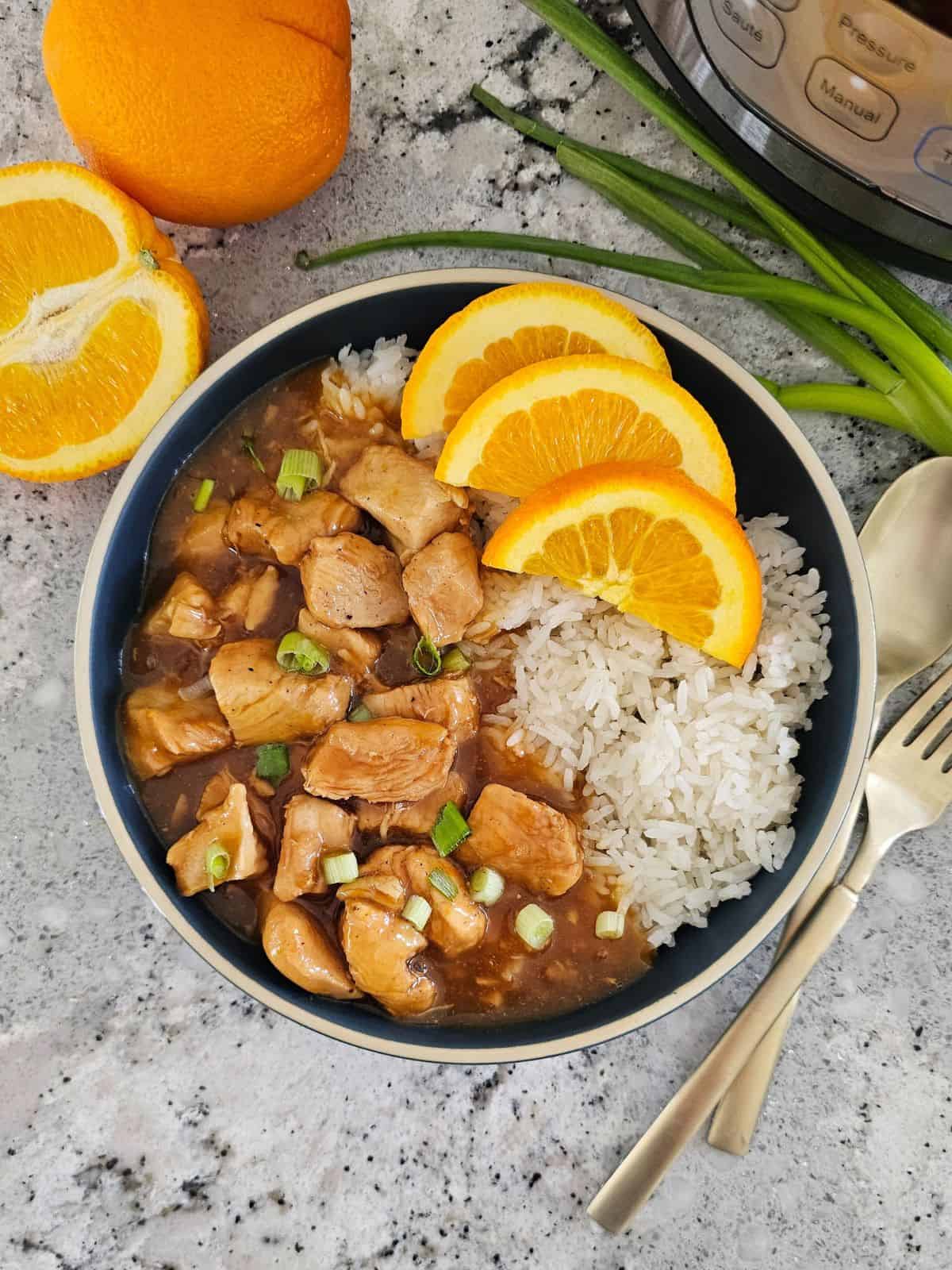 Instant Pot Orange Chicken served with rice on the side and the Instant Pot, and fresh oranges in the background.