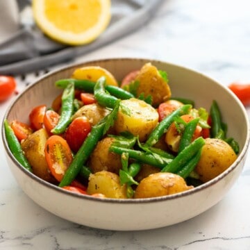Italian potato salad in bowl topped with fresh parsley with tomatoes in background.