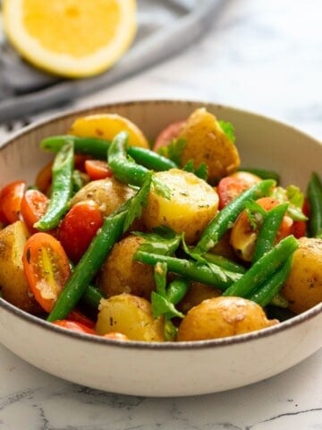 Italian potato salad in bowl topped with fresh parsley with tomatoes in background.