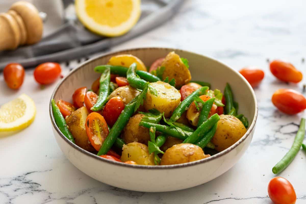 Italian potato salad in bowl topped with fresh parsley with lemon and tomatoes in background.