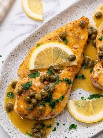 Chicken breast topped with lemon slices and piccata sauce on white platter.