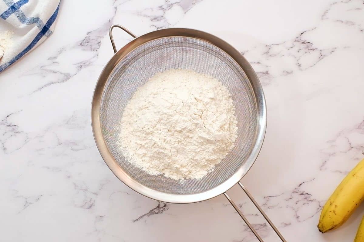 Metal sifter with flour, baking soda, and salt in it over a mixing bowl.