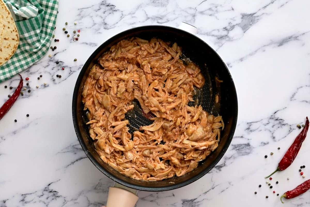 Shredded chicken tossed with buffalo sauce in saucepan.