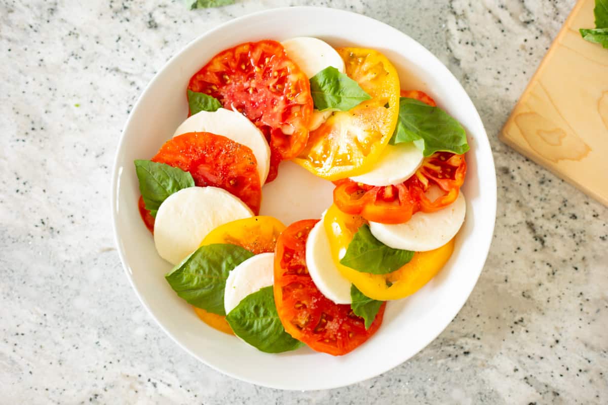 Alternating layers of tomatoes, mozzarella, and basil in shallow white serving platter.