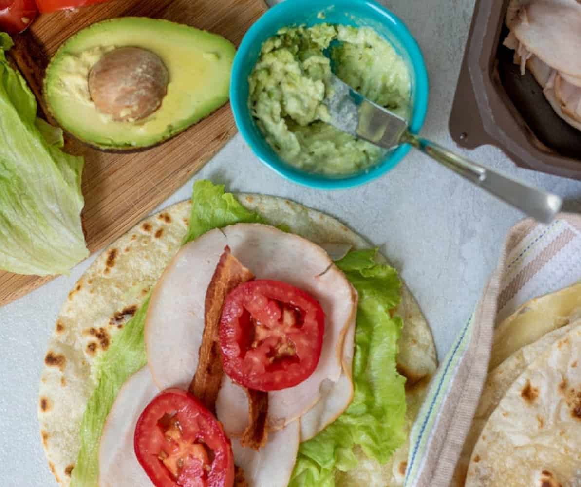 Tortilla with lettuce, turkey, tomatoes, and bacon next to mashed avocado.