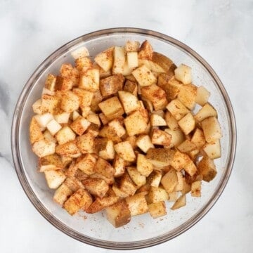 Cubed potatoes with dried seasonings in glass mixing bowl.
