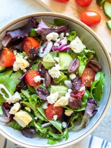 Greek Salad next to cutting board with fresh veggies.