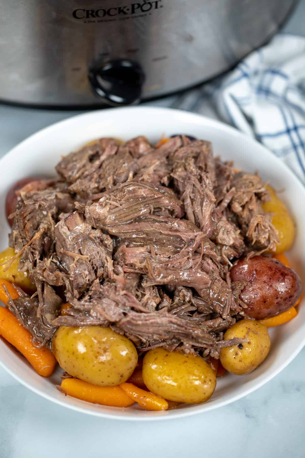 Shredded pot roast in white dish over potatoes and carrots next to crock pot.