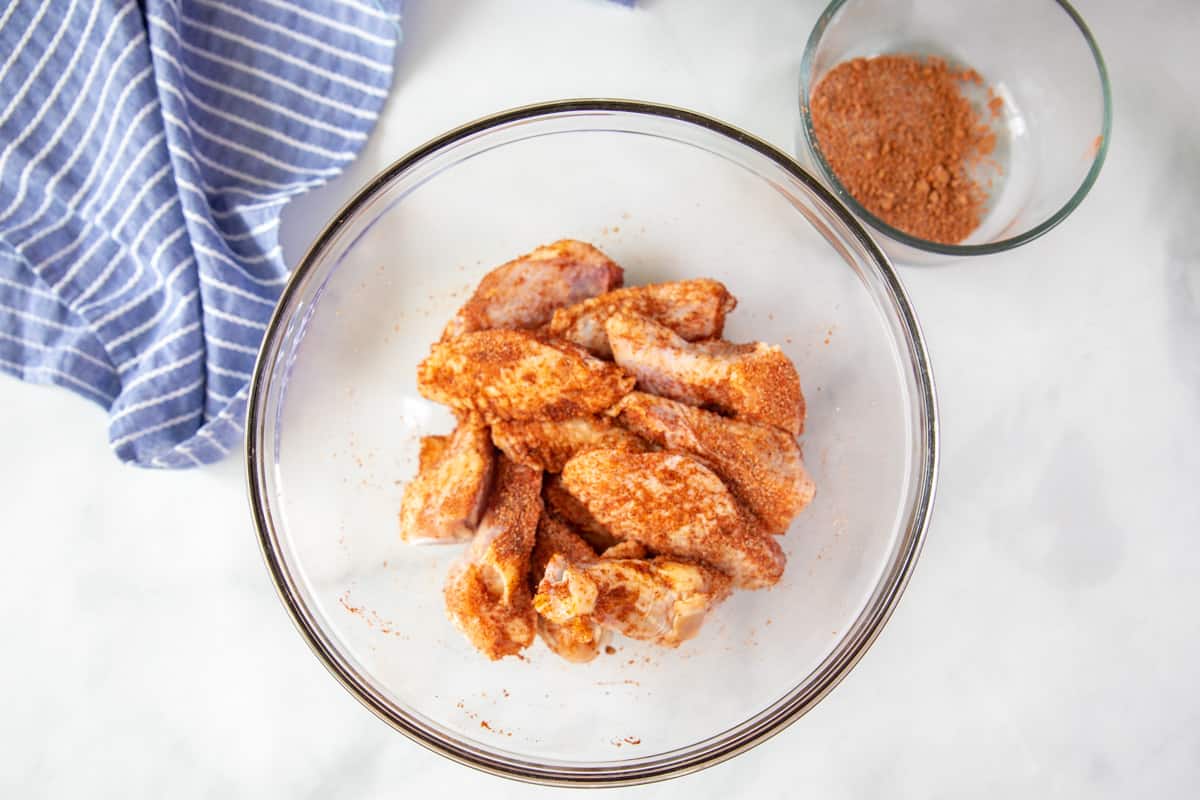 Chicken wings tossed with dry rub and oil in clear mixing bowl.