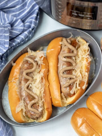 Two bratwurst in buns with sauerkraut and mustard next to instant pot.