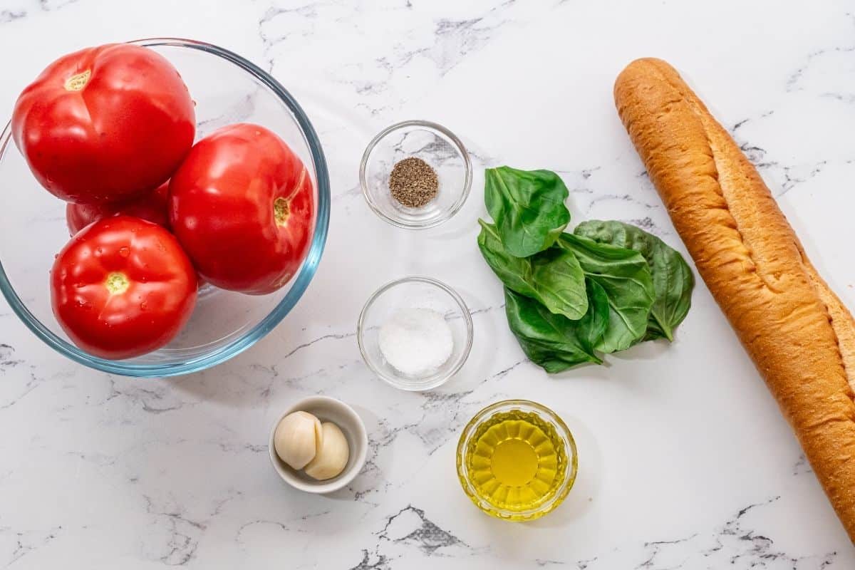 Fresh tomatoes, basil, garlic, olive oil, salt and baguette on counter. 