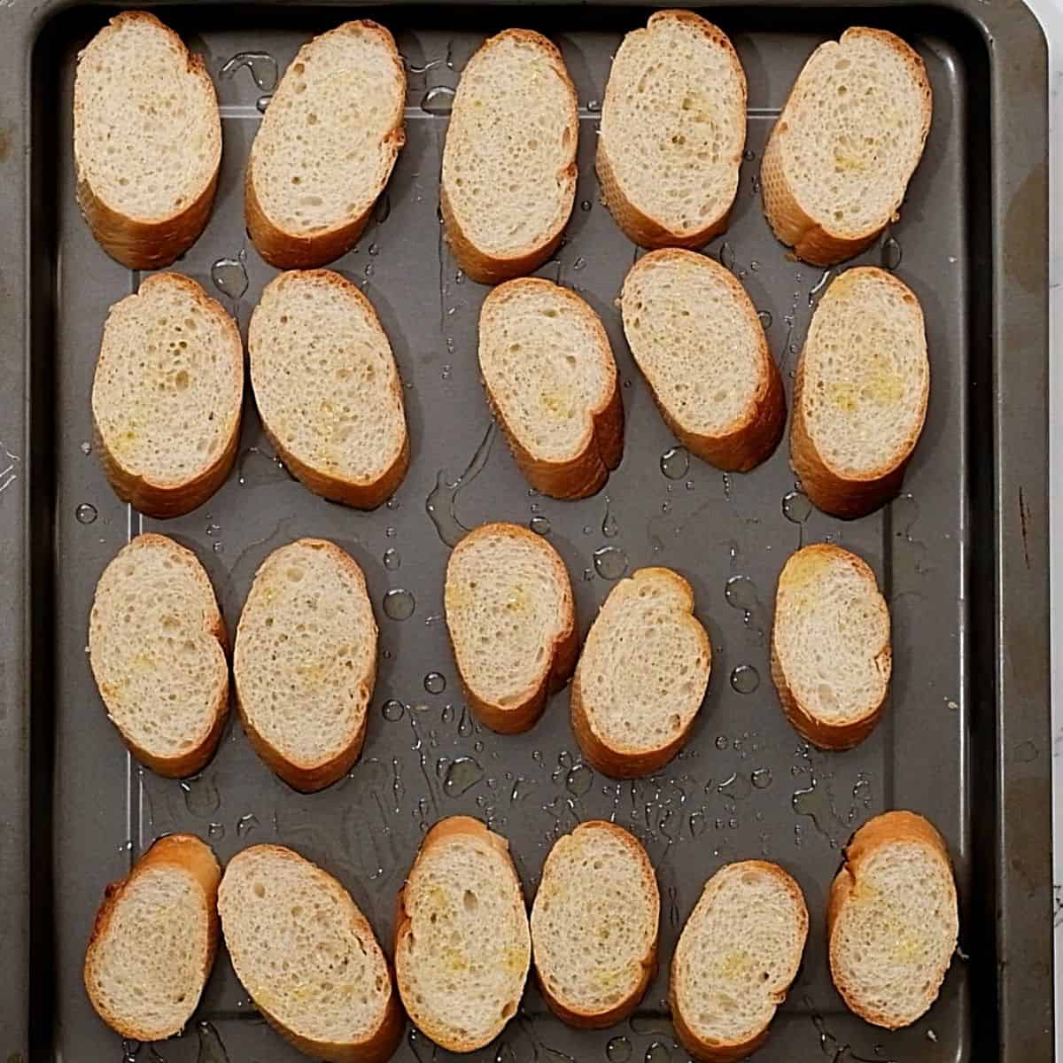 Sliced baguette on baking sheet with olive oil. 
