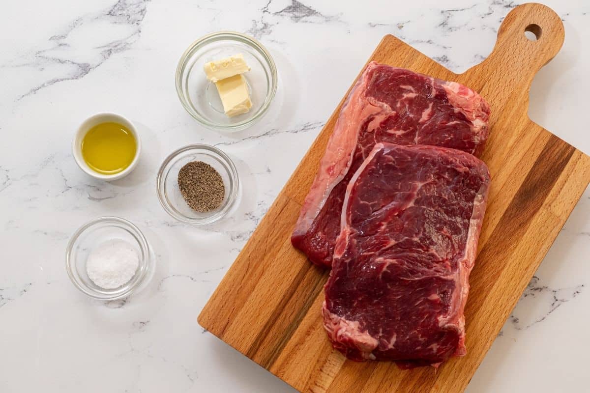 Raw ribeye steaks next to oil, salt, pepper, and butter. 