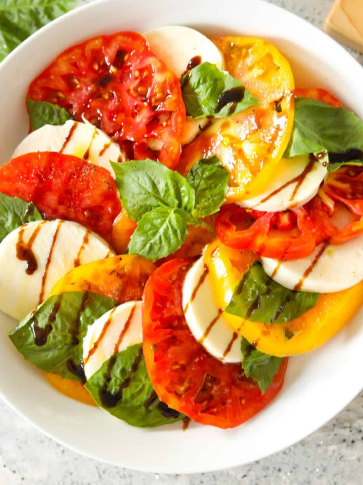 Alternating layers of tomatoes, mozzarella, and basil in shallow white serving platter topped with balsamic glaze.