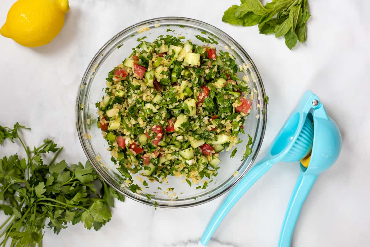Tabouli tossed together in clear bowl next to fresh lemon and parsley.