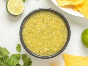 Easy Roasted Tomatillo Salsa Verde in black bowl with corn tortilla chips on the side with cilantro and limes in the background.