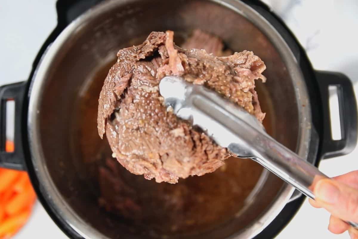Roast being removed from the inner pot of instant pot with metal tongs.