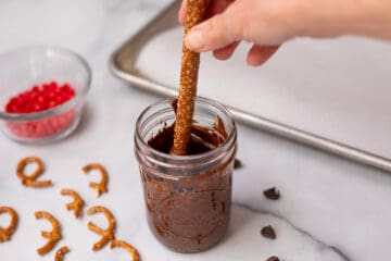 Pretzel rod being dipped half way into jar of melted chocolate.