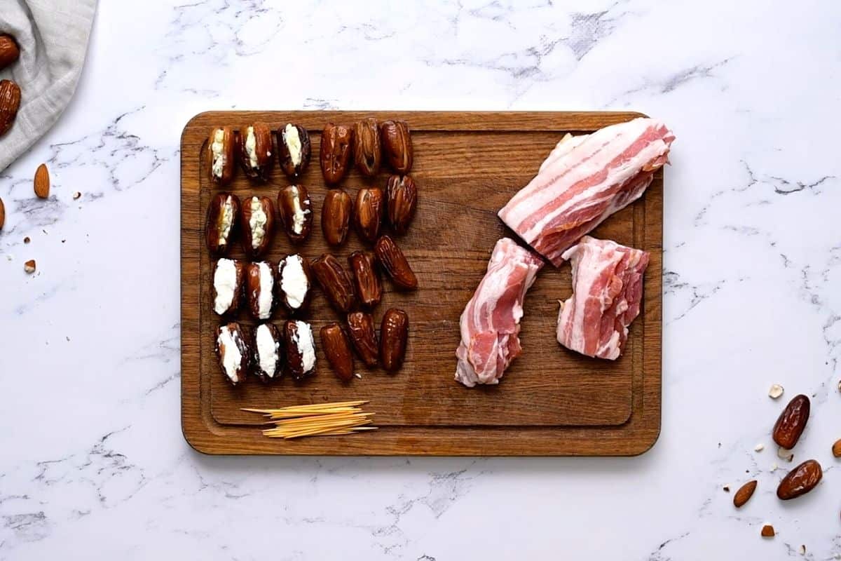 Dates stuffed with goat cheese, almonds, and blue cheese on cutting board next to bacon cut in half. 