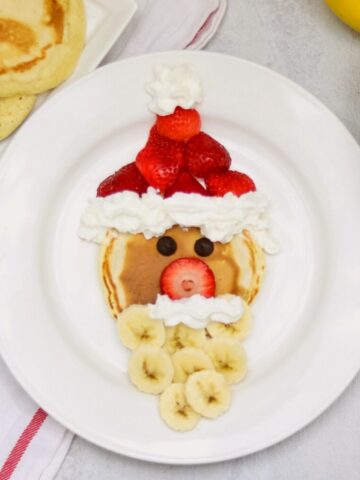 Santa made out of pancake, strawberries, and banana on white plate.