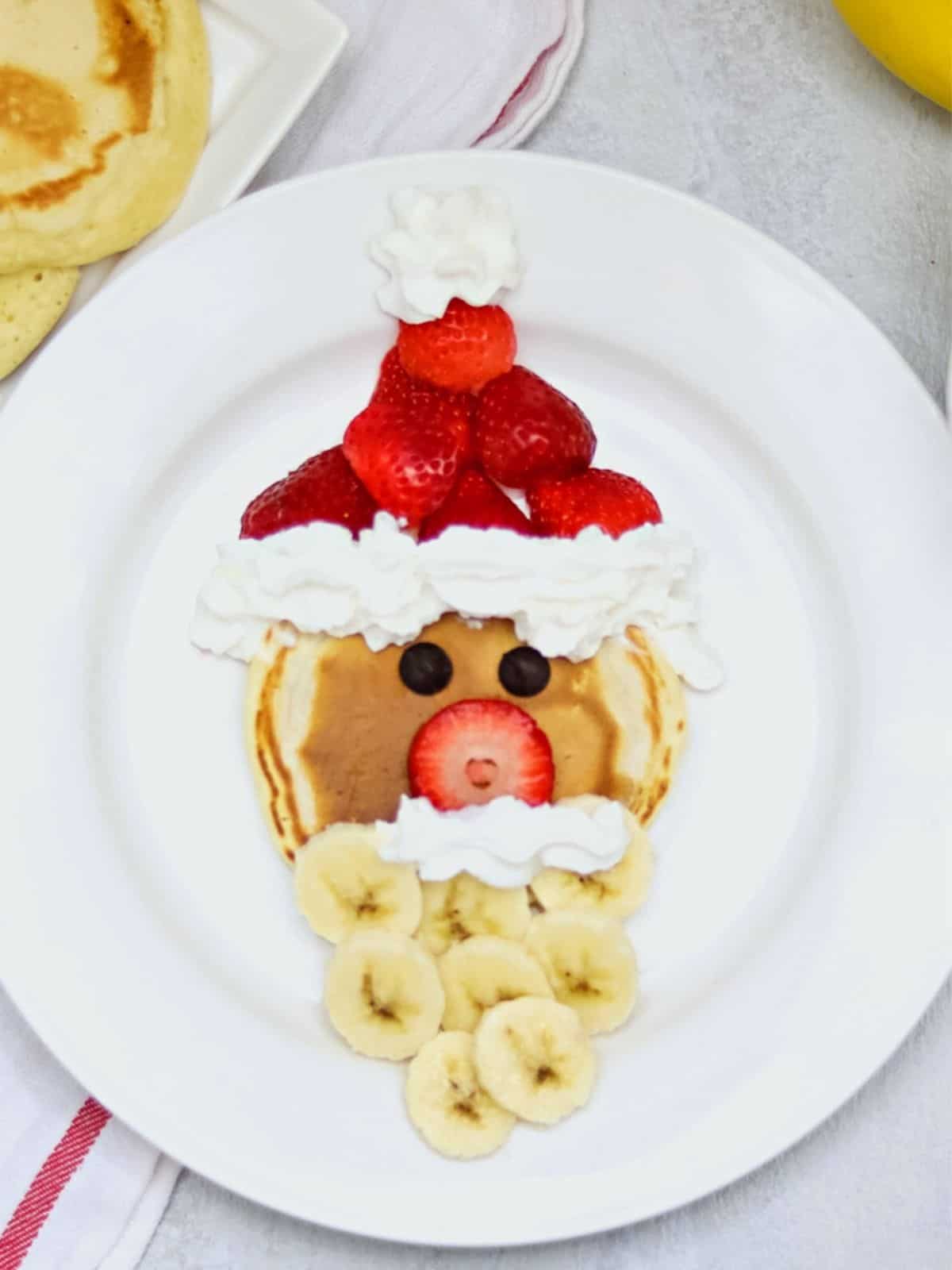 Santa shaped using a pancake, strawberries, whipped cream and bananas on white plate.