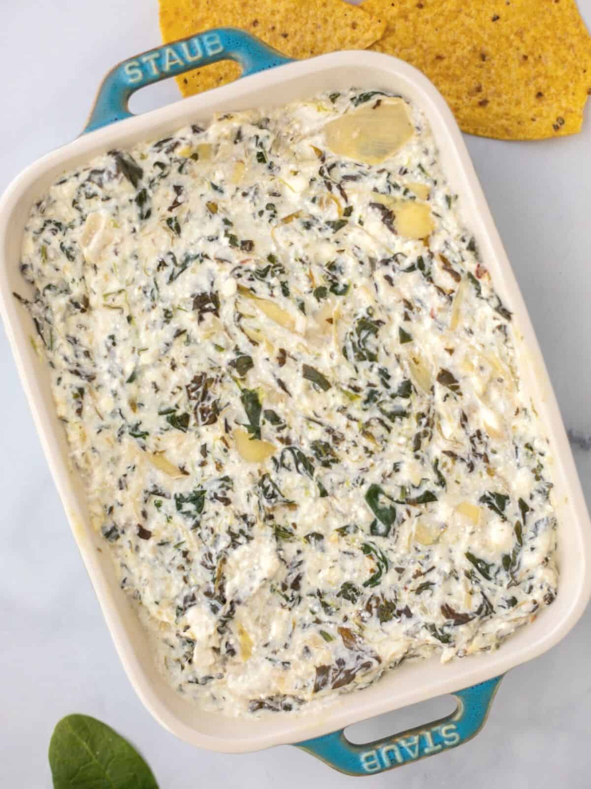 Spinach Artichoke Dip Spread out in blue casserole pan next to chips and fresh spinach.