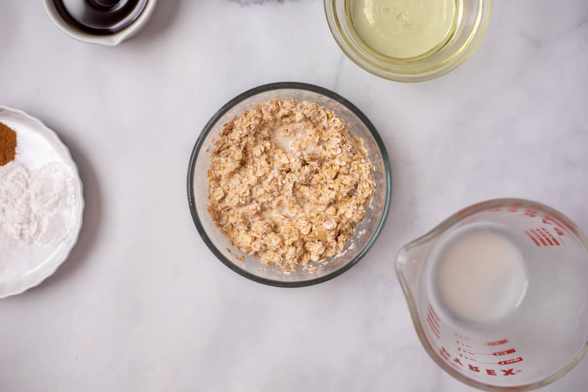 Oats and milk soaking in glass bowl next to other ingredients for muffins.