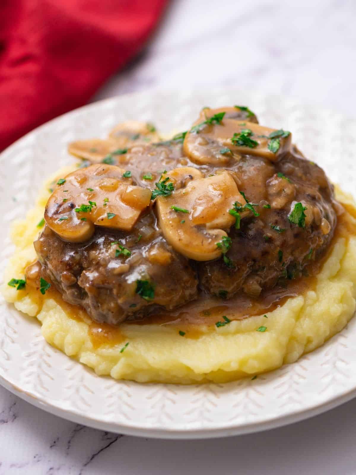 Chopped Steak with mushroom gravy served over mashed potatoes on white plate.